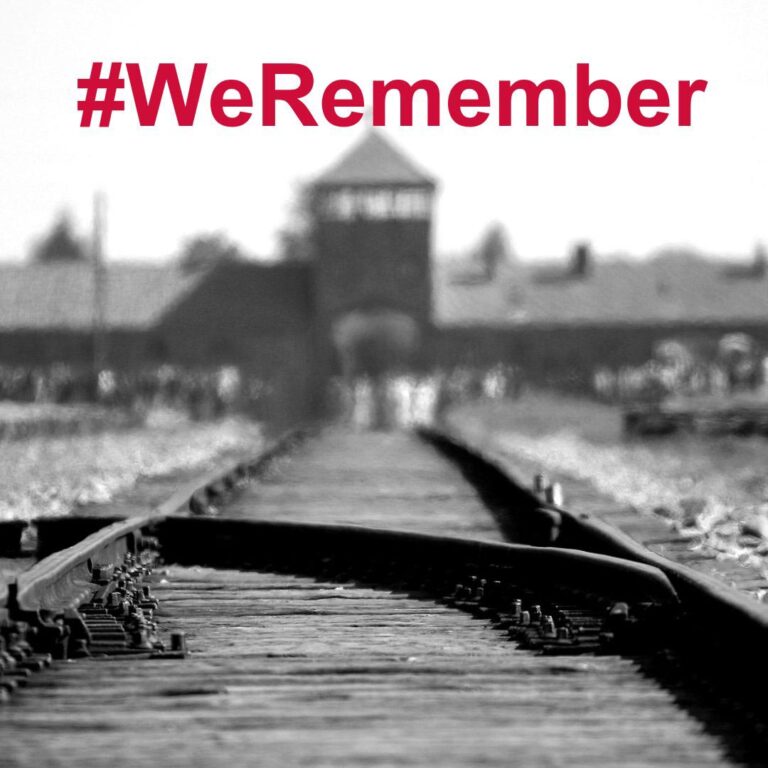 Schwarz-weiß-Foto mit Schienen im Vordergrund und dem Eingang zu einem KZ im Hintergrund